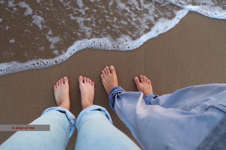 Two pairs of barefoot feet standing on sandy beach close to the water's edge, with gentle waves lapping at the shore.