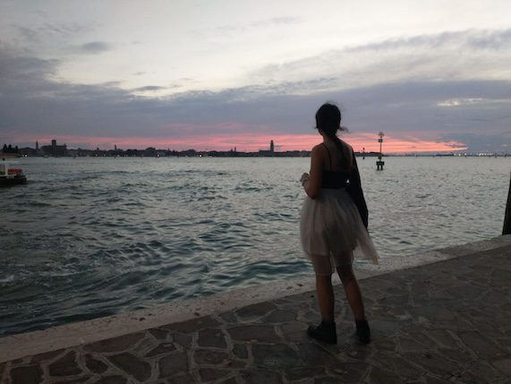 A person standing by the water's edge, facing away, with a city skyline in the background during sunset. This website is stories art and photos of an Iranian astrophysicist in Europe. she writes about how do Iranians feel about Israel for example